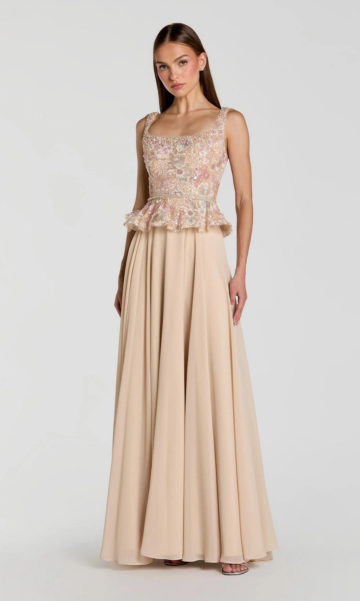 A woman wearing the Mac Duggal 21317 long formal dress, an elegant sleeveless, floor-length beige a-line chiffon evening gown featuring a floral embroidered bodice and peplum waistline, stands against a plain light background.